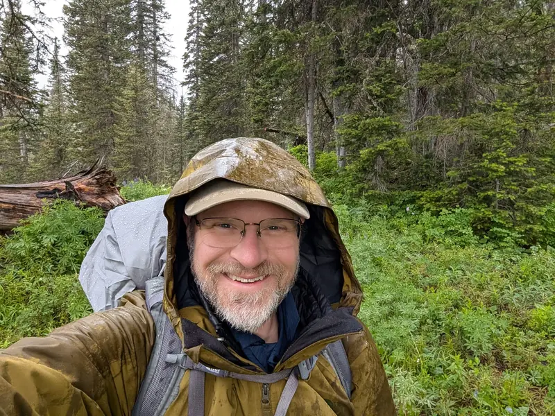Selfie in a rain-soaked jacket and hood with a covered backpack among evergreens